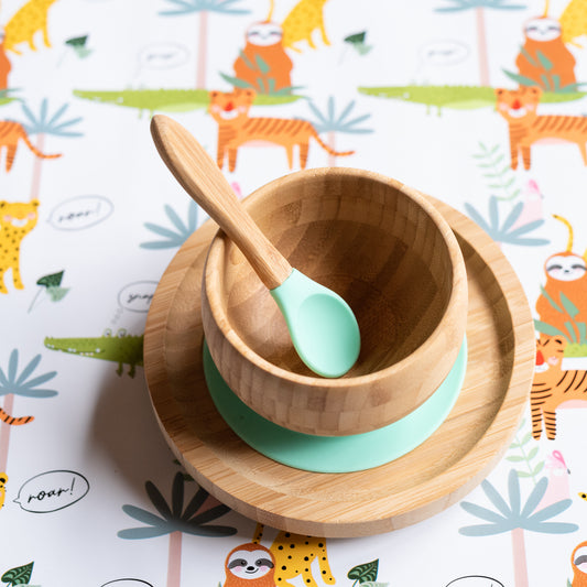 A wooden baby bowl with a mint blue silicone liner and matching spoon, set on a round wooden plate over a white background with a colorful animal print.