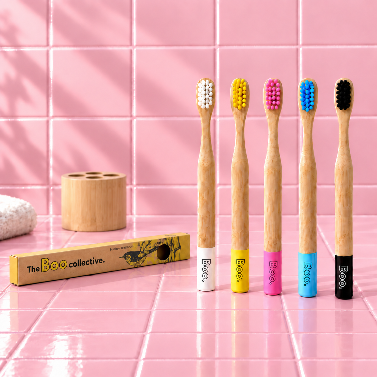 Four bamboo toothbrushes with colorful bristles and branded bases are lined up on a pink tiled surface, with a matching toothbrush box and wooden holder in the background.