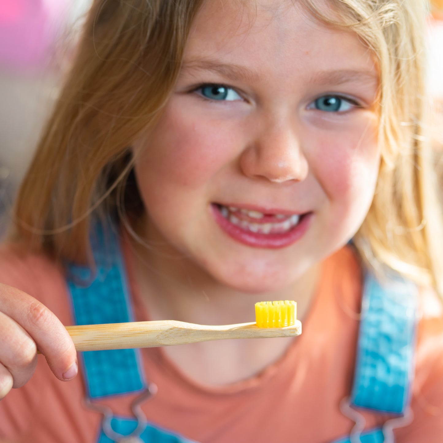 A young girl with blonde hair smiling while holding a bamboo toothbrush with yellow bristles, wearing a peach shirt and blue denim overalls.