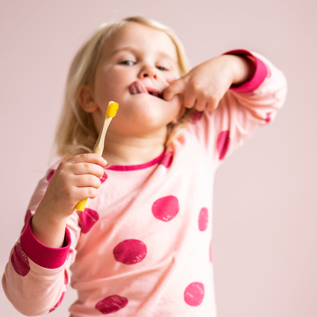 A young child wearing pink pajamas with red polka dots holds a yellow toothbrush while stretching their tongue, set against a solid light pink background.