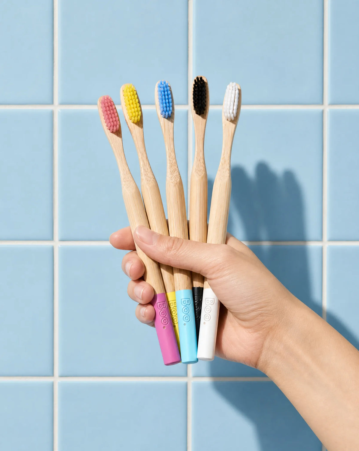 A hand holding five bamboo toothbrushes with colorful bristles and pastel-colored handles, set against a light blue tiled wall.