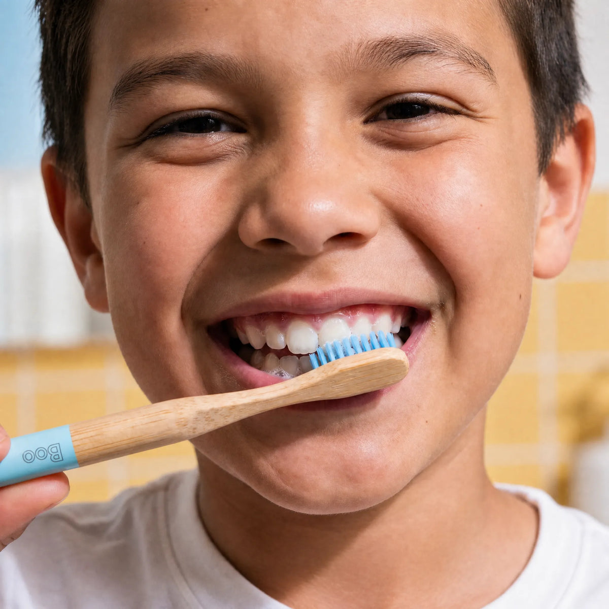 A close-up lifestyle shot of a smiling young boy brushing his teeth with a bamboo toothbrush featuring a light blue handle and blue bristles.