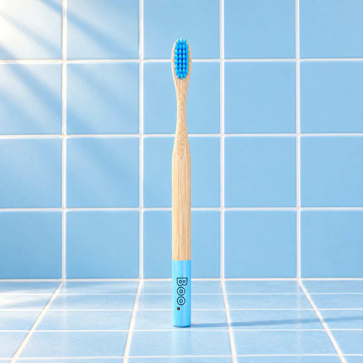 Boo adult bamboo toothbrush with blue bristles and a blue end cap, standing on blue tiles