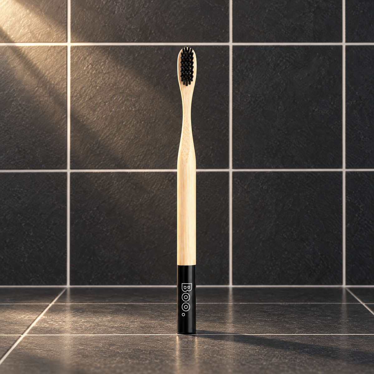 Boo adult bamboo toothbrush with black bristles and a black end cap, standing on charcoal tiles