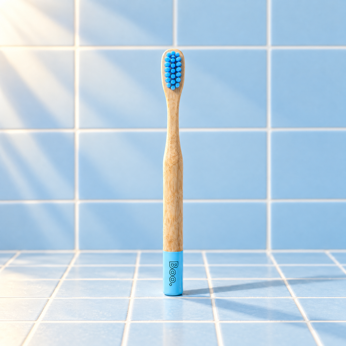 Boo kids bamboo toothbrush with blue bristles and a blue end cap, standing on blue tiles