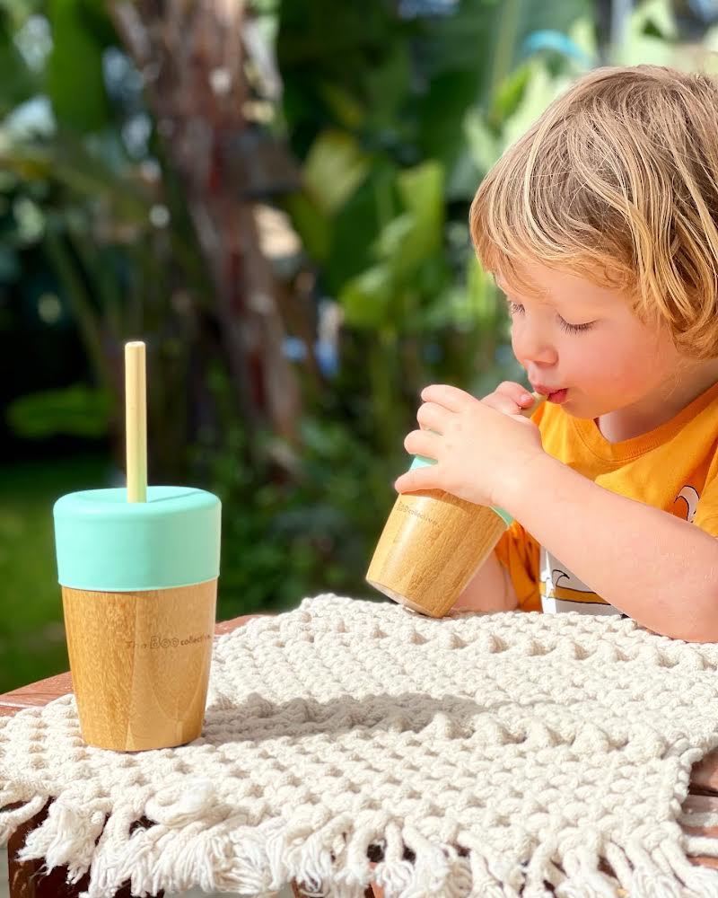 Child drinking from a wooden cup with a straw, outdoors on a sunny day.